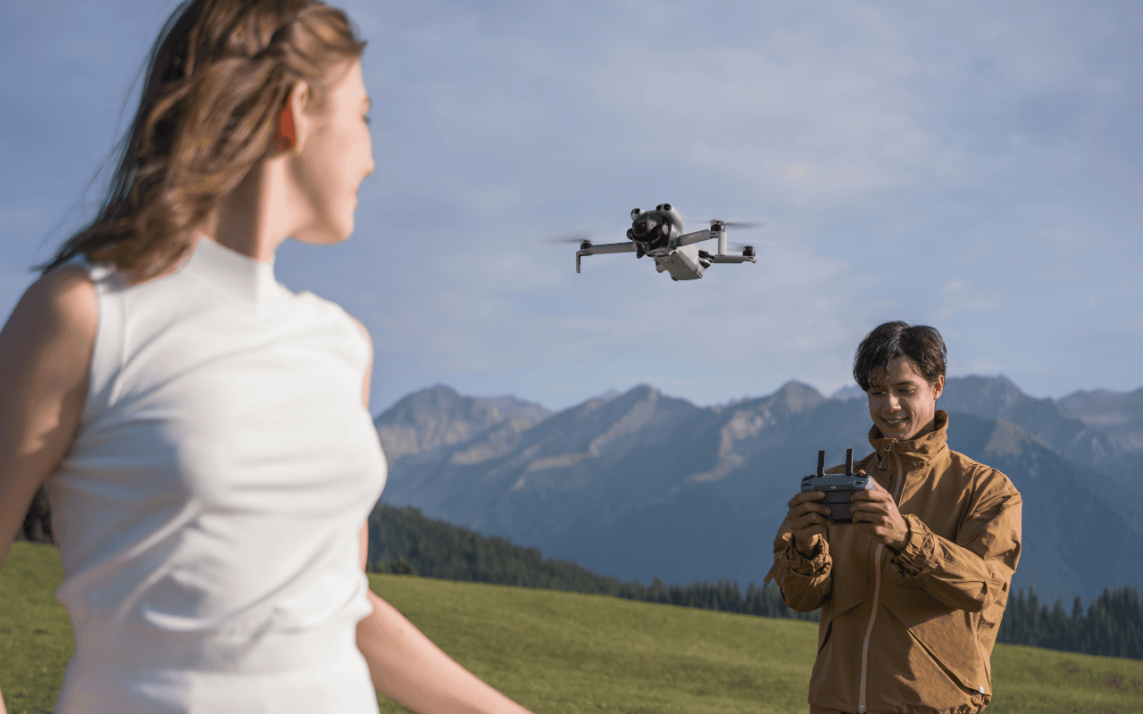 Auf dem Foto ist die DJI Mini 5 Pro im Flug zu sehen. Der Pilot ist zudem lächelnd im Hintergrund. Das Panorama im Hintergrund zeigt eine Berglandschaft.
